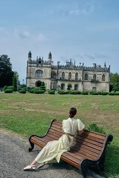Темнокожая девушка в светлом платье отдыхает на скамейке перед старинным замком