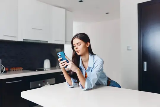 Молодая женщина с телефоном облокотилась на стол на светлой кухне