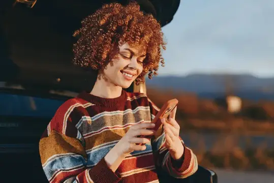 Кудрявая девушка улыбается, глядя в телефон на закате