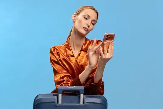 Светловолосая женщина в оранжевой рубашке смотрит в телефон у чемодана