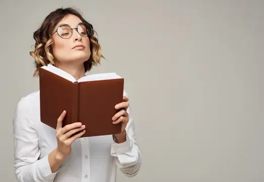 Женщина с коричневым блокнотом на сером фоне, на лице у неё очки