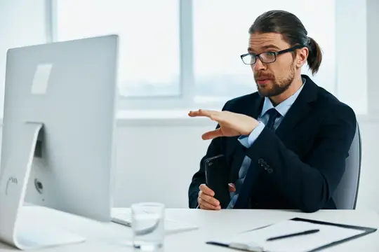 Бородатый мужчина в очках жестикулирует перед монитором в офисе