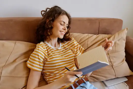 Улыбающаяся кудрявая девушка читает книгу на диване с подушками