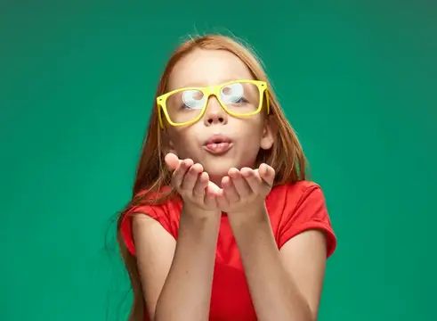Рыжеволосая девочка в жёлтых очках посылает воздушный поцелуй на зелёном фоне