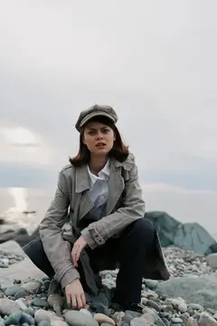 Портрет молодой женщины в ретро-пальто и шляпе, сидящей на корточках на каменном пляже у моря под пасмурным небом