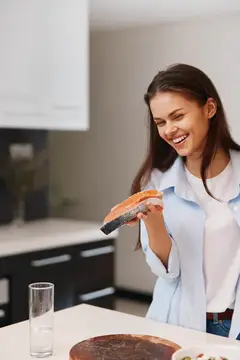 Улыбающаяся темноволосая девушка держит сэндвич на светлой кухне