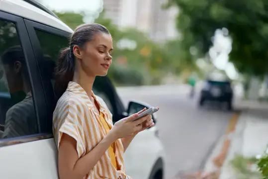 Молодая женщина со смартфоном стоит у белой машины на улице