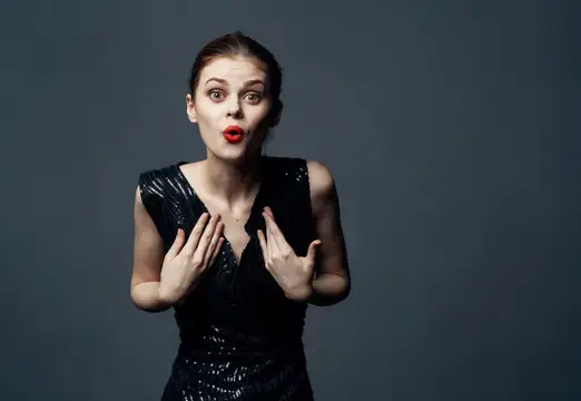 Удивлённая женщина в чёрном платье прижимает руки к груди