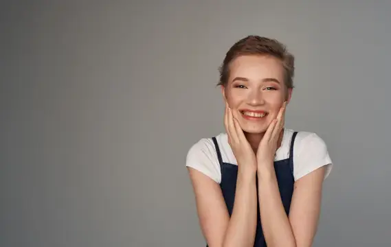 Улыбающаяся девушка с короткой стрижкой держит руки у лица на сером фоне