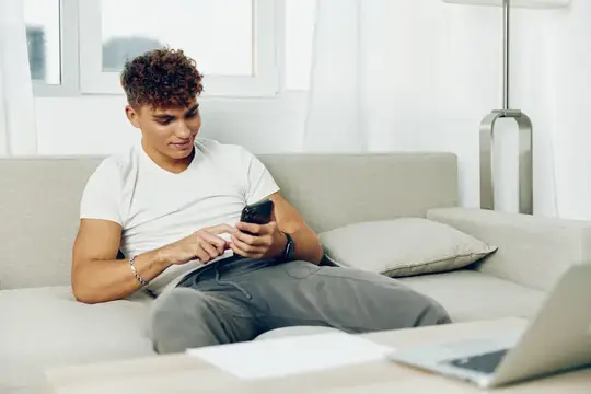 Кудрявый парень в белой футболке сидит на диване со смартфоном