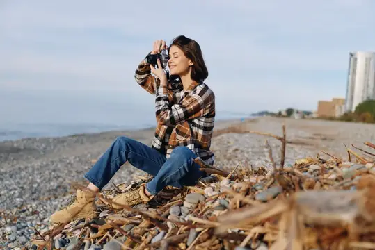 Молодая женщина с пленочным фотоаппаратом сидит на каменистом берегу