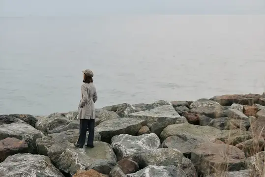 Женщина в плаще и кепке стоит на камнях у туманного моря