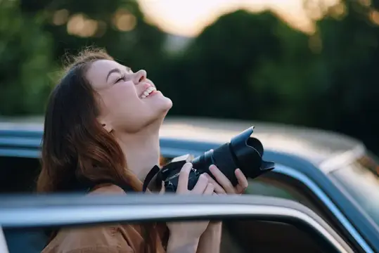 Смеющаяся девушка с фотокамерой выглядывает из окна автомобиля
