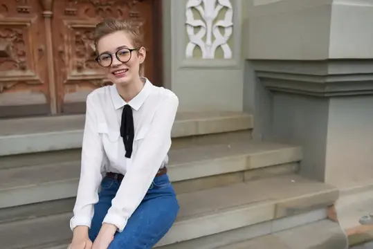 Улыбчивая девушка в очках сидит на каменных ступенях у старинного здания