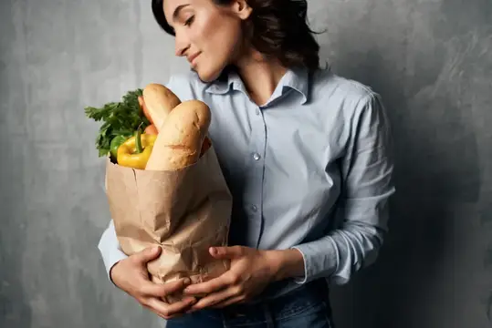 Весёлая брюнетка с пакетом продуктов, в голубой рубашке, на фоне овощей и тёмного фона