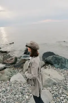 Молодая женщина в кепке и плаще стоит на каменистом берегу моря