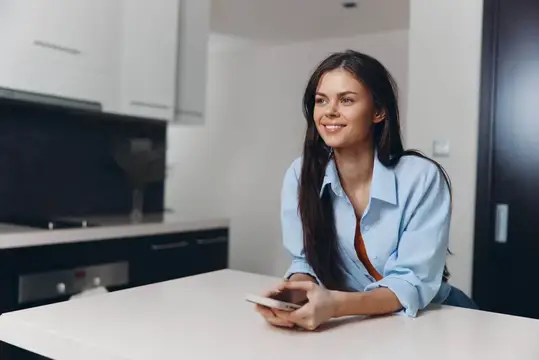 Улыбающаяся темноволосая девушка с телефоном на светлой кухне