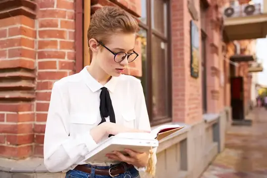 Молодая девушка в очках читает книгу у кирпичного здания