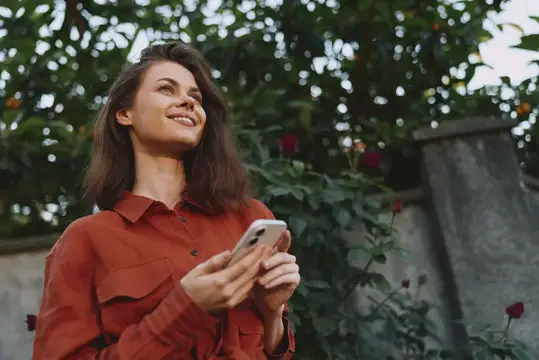 Взрослая женщина среди зелени поднимает взгляд с мягкой улыбкой, листая смартфон