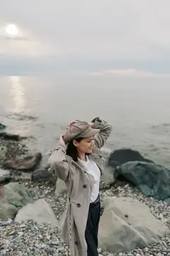 Молодая женщина в плаще и кепке стоит на каменистом берегу моря