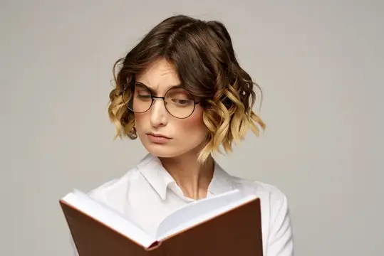 Молодая женщина в очках с книгой сосредоточенно хмурится