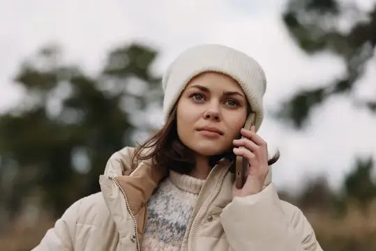 Портрет женщины в теплой куртке и вязаной шапке, разговаривающей по мобильному устройству, уличная обстановка с пасмурным небом, сезонная мода и откровенный момент