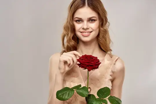 Улыбающаяся рыжеволосая девушка держит красную розу на сером фоне