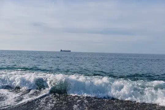 Волна разбивается о гальку на фоне моря и сухогруза