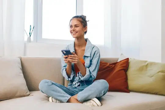 Улыбающаяся девушка со смартфоном сидит по-турецки на диване