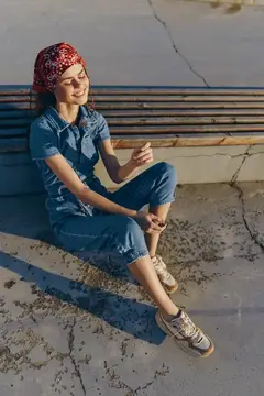 Девушка в джинсовом комбинезоне и красной бандане сидит на скамейке