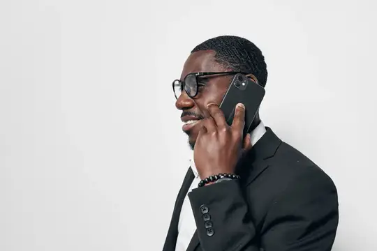 Афроамериканский бизнесмен с темной кожей в официальном черном костюме и очках разговаривает по мобильному телефону