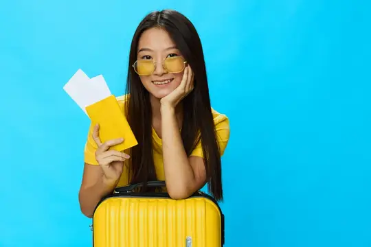 Улыбающаяся азиатская девушка с билетами и желтым чемоданом на синем фоне