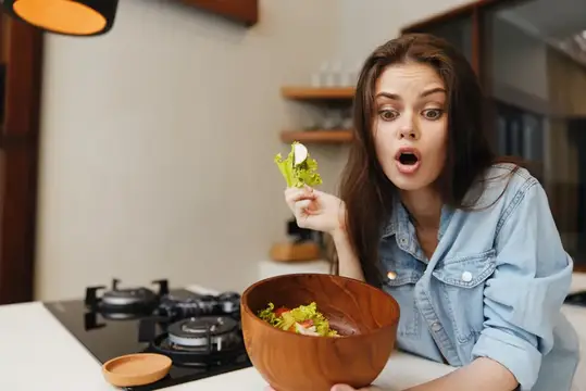 Удивлённая девушка с листом салата над миской на кухне