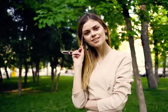 Молодая женщина с очками в руке улыбается в летнем парке