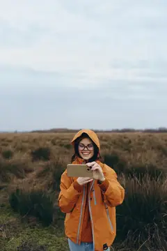 Улыбающаяся девушка в оранжевой куртке фотографирует на телефон осеннее поле