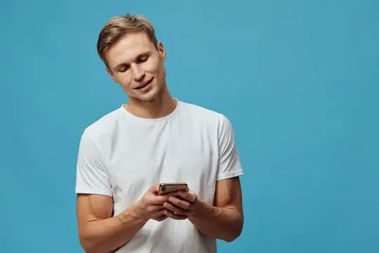 Молодой блондин в белой футболке смотрит в телефон на синем фоне