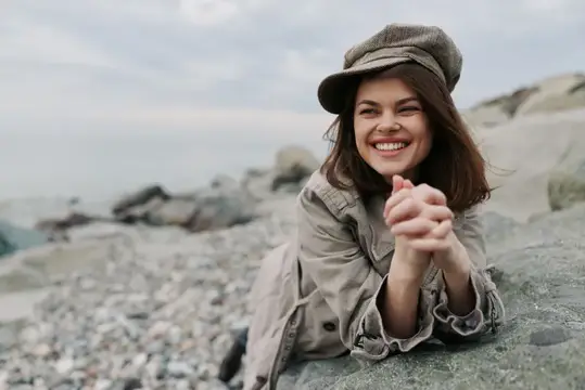 Улыбающаяся девушка в кепке лежит на каменистом берегу