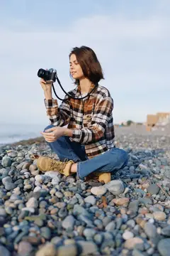 Непринужденный фотограф на открытом воздухе на каменистом берегу, объектив готов, документирует береговую линию камерой, клетчатая рубашка, джинсы и спокойное выражение лица на фоне морского бриза