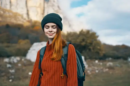 Рыжеволосая девушка в шапке и свитере с рюкзаком на фоне гор