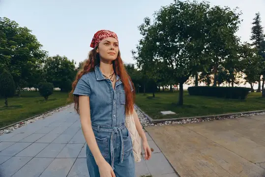 Рыжеволосая девушка в джинсовом комбинезоне и бандане гуляет по парку