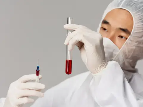 Азиатский лаборант в защитном костюме рассматривает пробирку с кровью