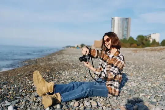 Улыбающаяся девушка с фотоаппаратом сидит на галечном берегу у моря