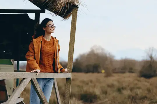 Улыбающаяся девушка в очках и оранжевой куртке у деревянной беседки