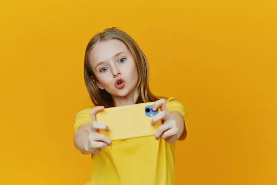 Светловолосая девочка с удивлением фотографирует на желтый смартфон