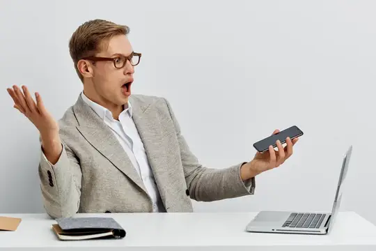 Удивлённый мужчина в очках смотрит на смартфон за рабочим столом