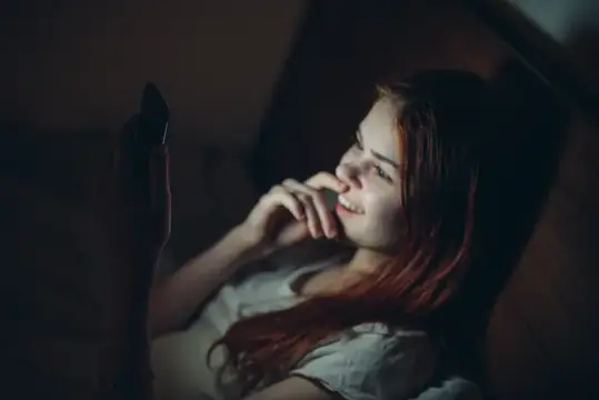 Рыжеволосая девушка лежит в темноте со смартфоном