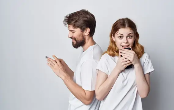 Удивлённая девушка с телефоном стоит спиной к бородатому мужчине