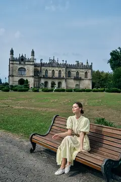 Молодая женщина в светлом платье отдыхает на скамейке перед старинным замком