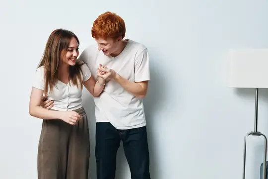 Рыжеволосый парень и девушка смеются, держась за руки у стены