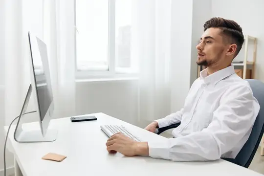 Молодой мужчина в белой рубашке работает за компьютером у окна